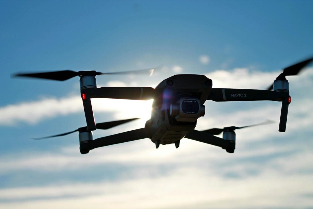 A Mavic 2 drone captured in flight with the sun shining through against a clear blue sky.