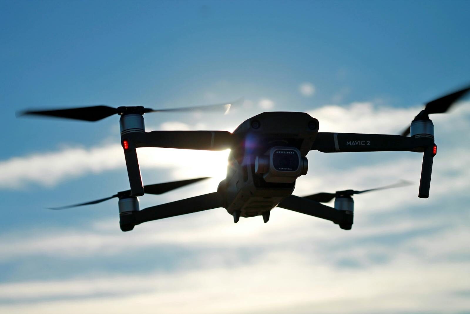 A Mavic 2 drone captured in flight with the sun shining through against a clear blue sky.